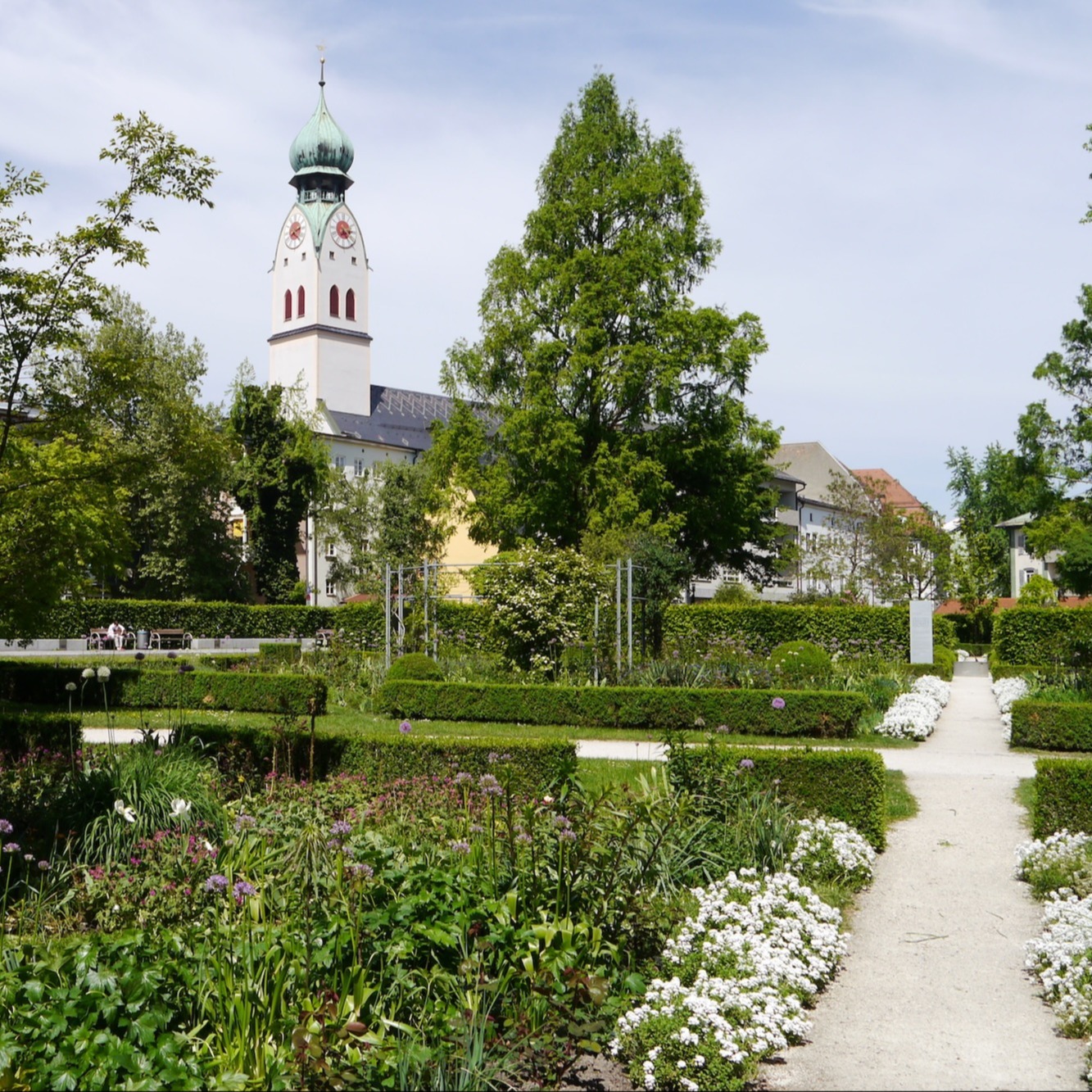 Rosenheim in Bayern: Grüner Garten mit Blumenbeeten, Büschen und Wegen, im Hintergrund ein Kirchturm.