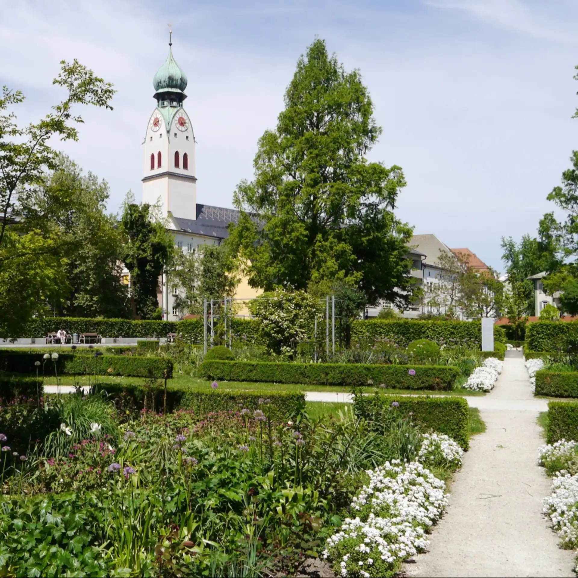 Rosenheim in Bayern: Grüner Garten mit Blumenbeeten, Büschen und Wegen, im Hintergrund ein Kirchturm.