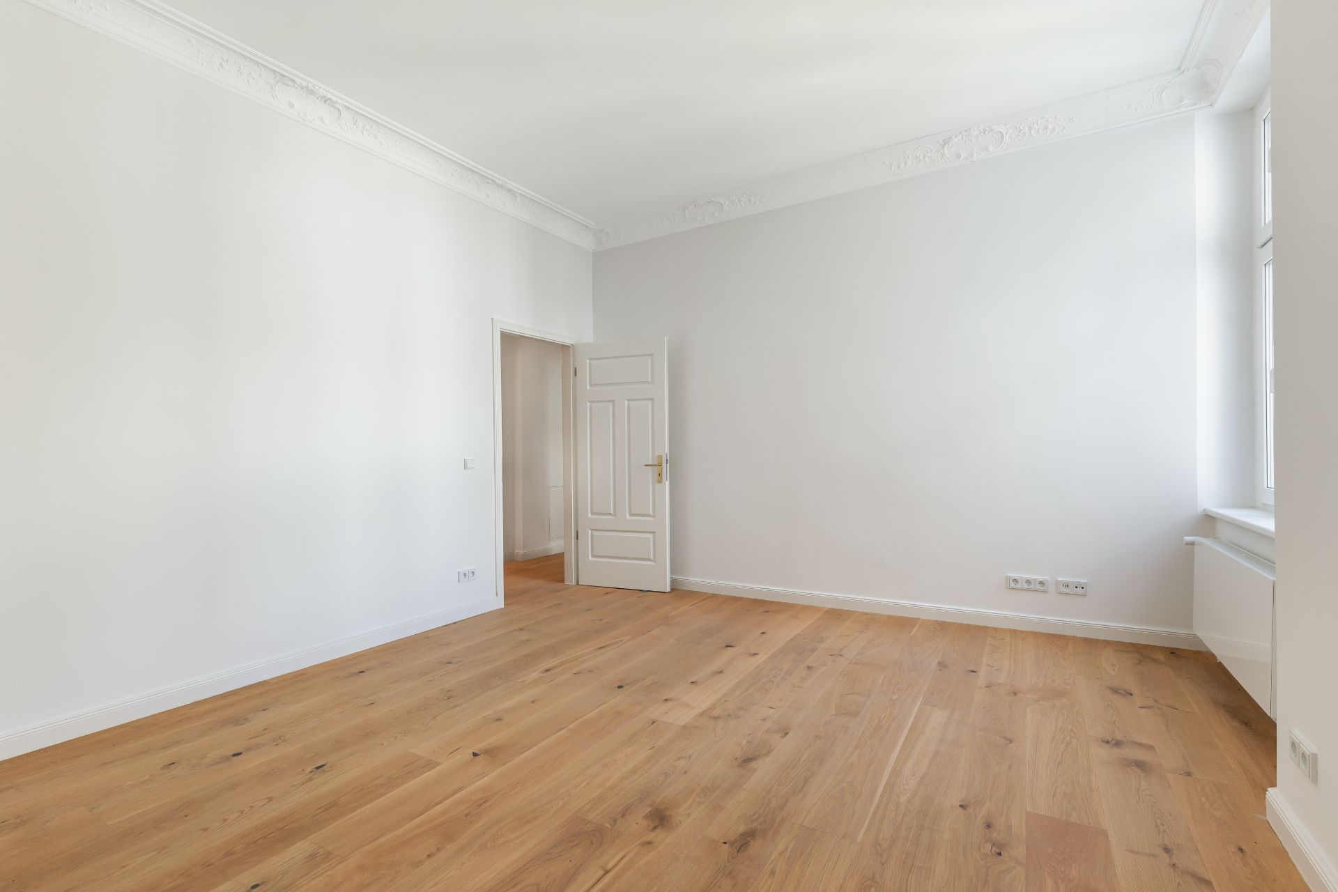 Leerer Raum mit hellem Holzboden, weißen Wänden, Tür und großem Fenster.