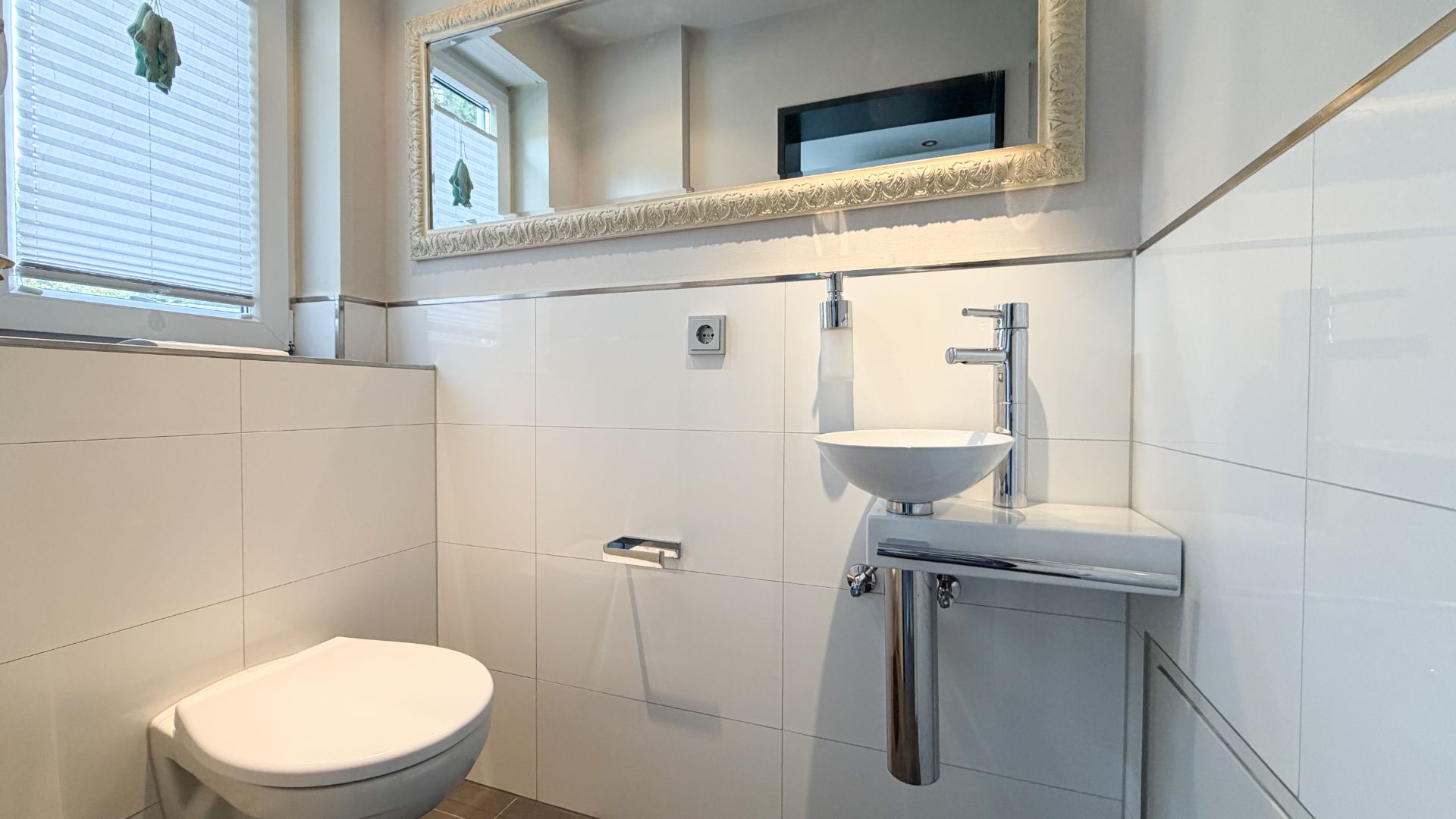 Modernes, weiß gefliestes Bad mit Wandtoilette, kleinem Waschbecken und großem Spiegel.