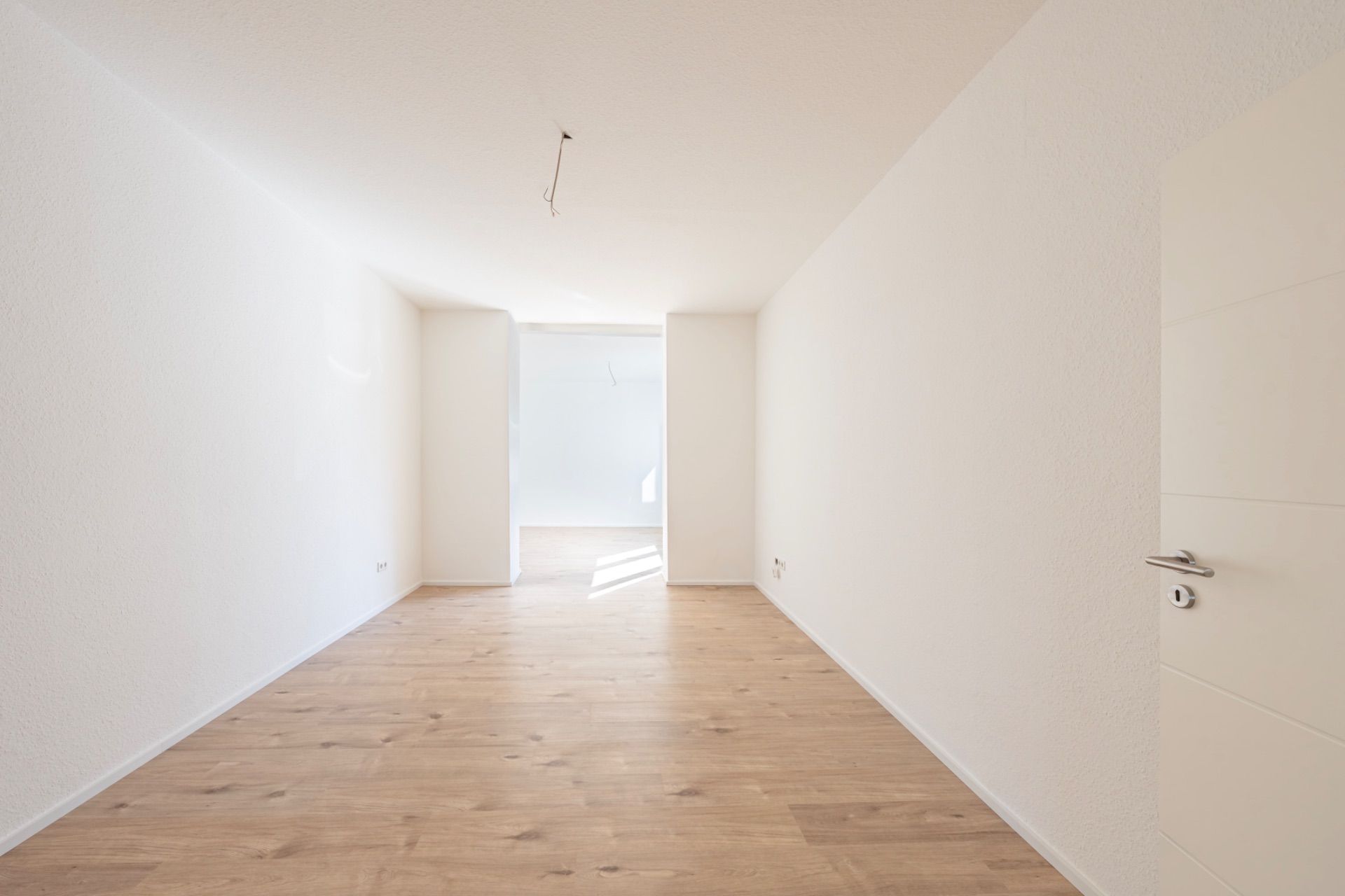 Leerer, heller Raum mit Holzfußboden und weißen Wänden, Tür rechts im Vordergrund.