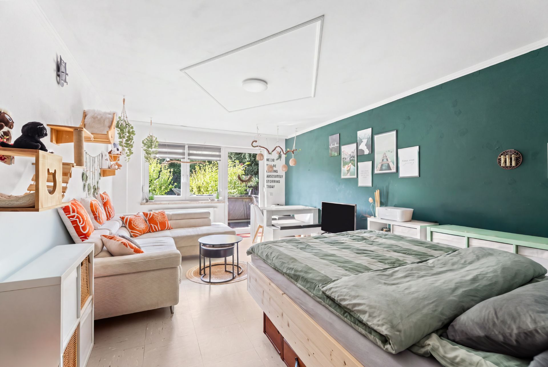Helles Wohn- und Schlafzimmer mit Bett, Sofa, grüne Wand und Fenster mit Blick ins Grüne.