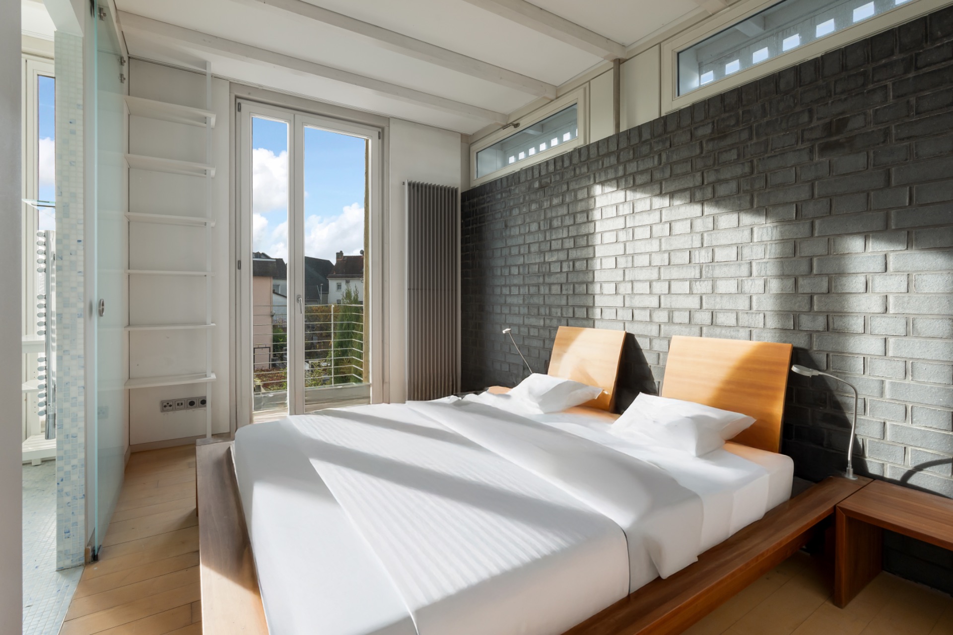 Modernes Schlafzimmer mit großem Bett, Holzboden und grauer Backsteinwand, viel Tageslicht.