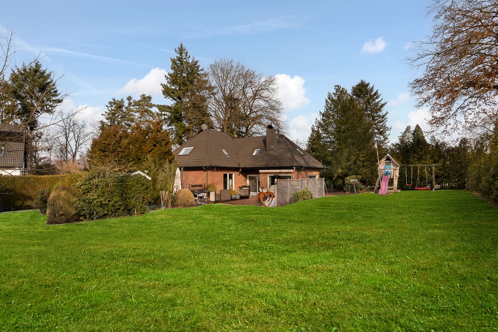 Haus mit großem Garten, Terrasse und Kinderspielplatz mit Rutsche und Schaukel, umgeben von Bäumen.