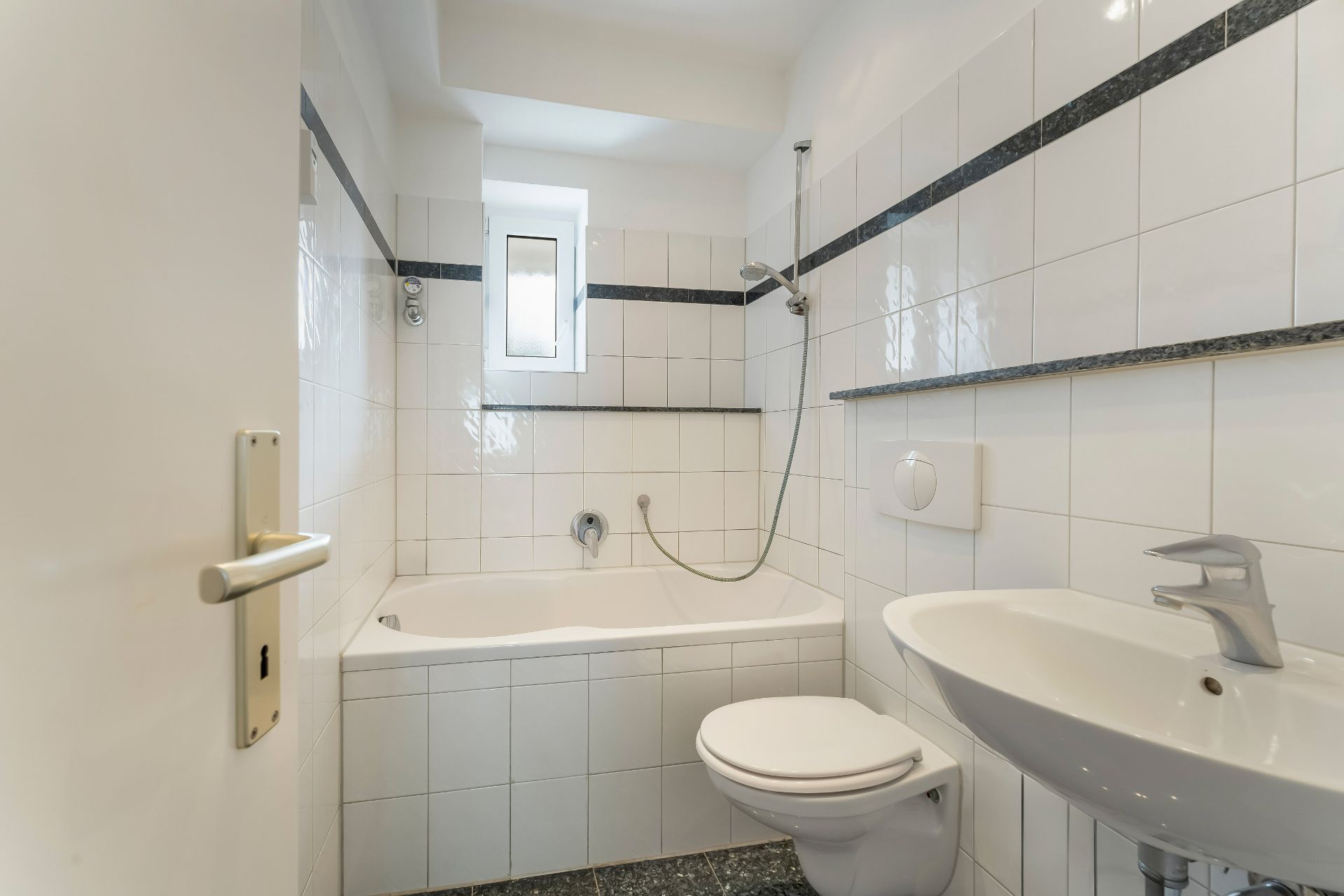 Helles Badezimmer mit Badewanne, Wandtoilette, Waschbecken und kleinem Fenster.