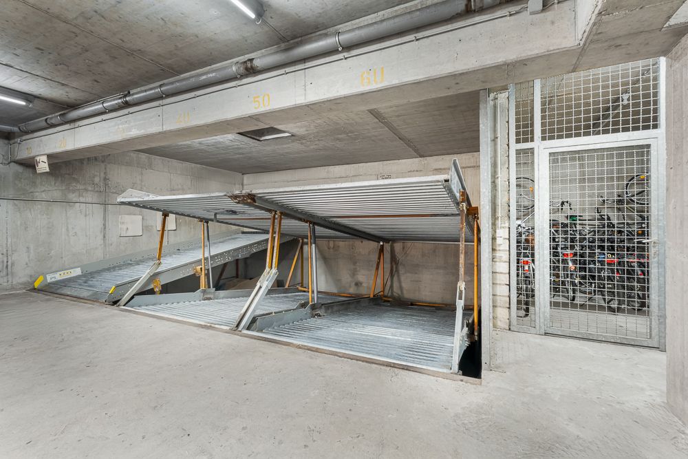 Mehrstöckige Parkplätze aus Metall in einer Tiefgarage, daneben ein Gitterraum mit Fahrrädern.