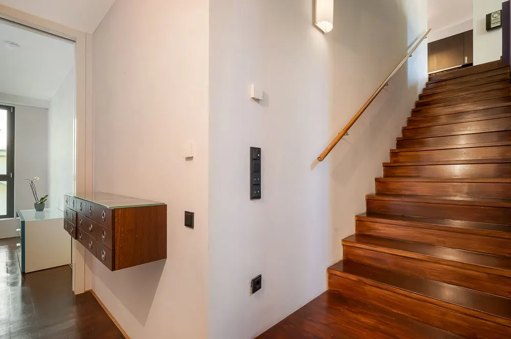 Holztreppe mit Handlauf neben Flur mit Wandkonsole und Blick in ein Zimmer mit Fenster.