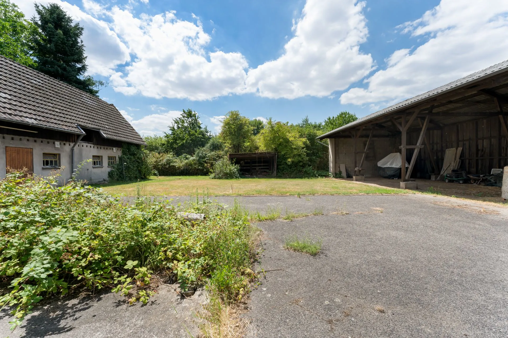 Hof mit Wohnhaus links, offenem Schuppen rechts und viel Grün im Hintergrund.