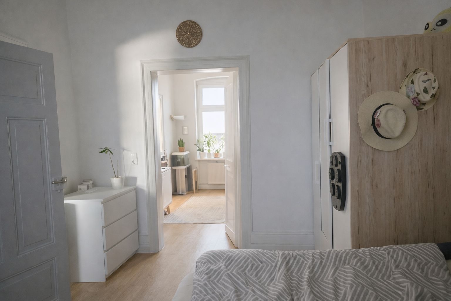 Helles Schlafzimmer mit Bett, Kommode, Schrank und Blick in einen weiteren Raum mit Fenster.