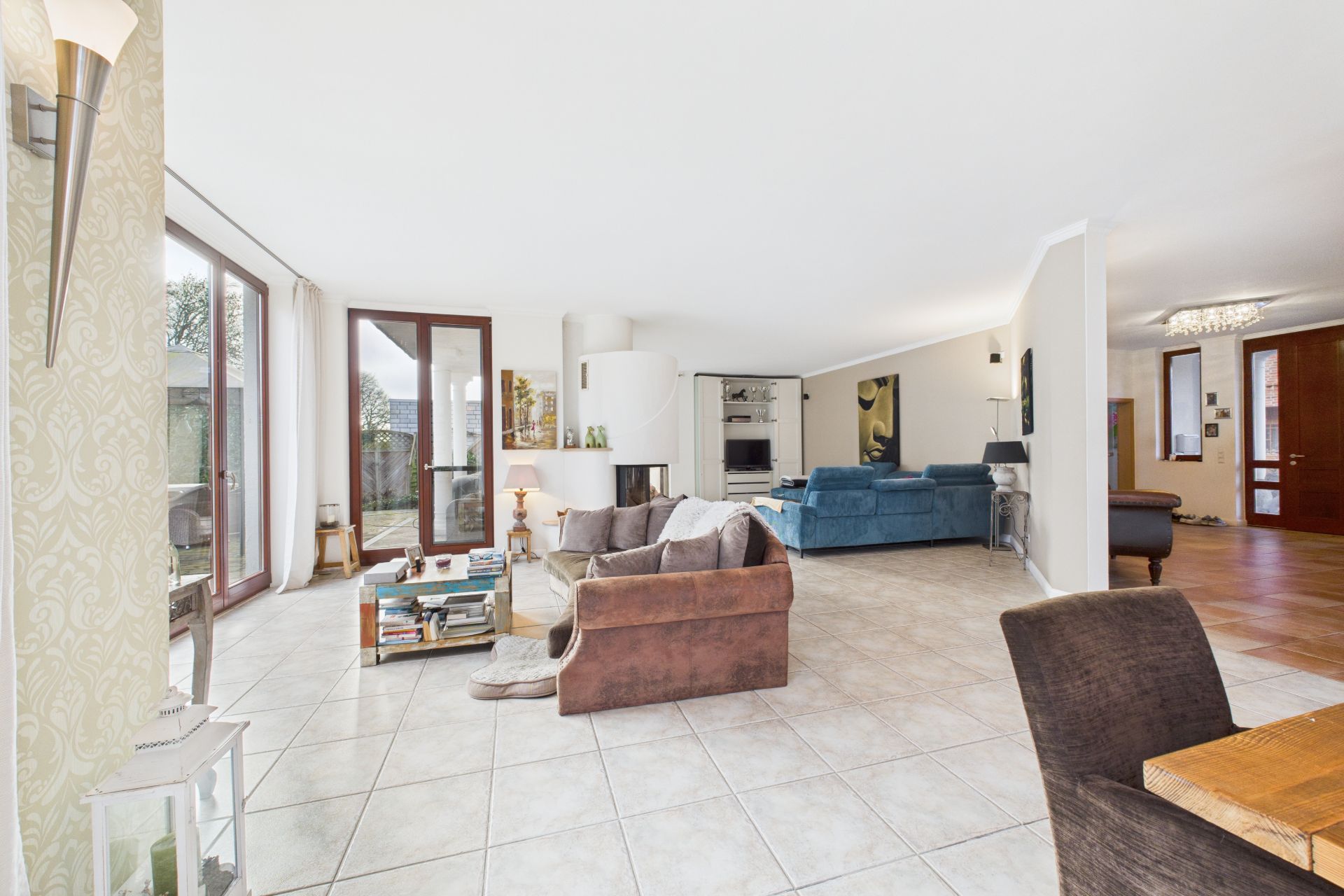Helles Wohnzimmer mit Sofas, Couchtisch, Kamin und großen Fenstern, Fliesenboden.