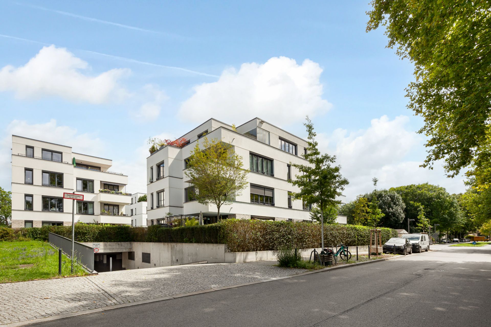 Moderne Wohngebäude mit weißen Fassaden an einer ruhigen Straße, umgeben von Bäumen und Autos.