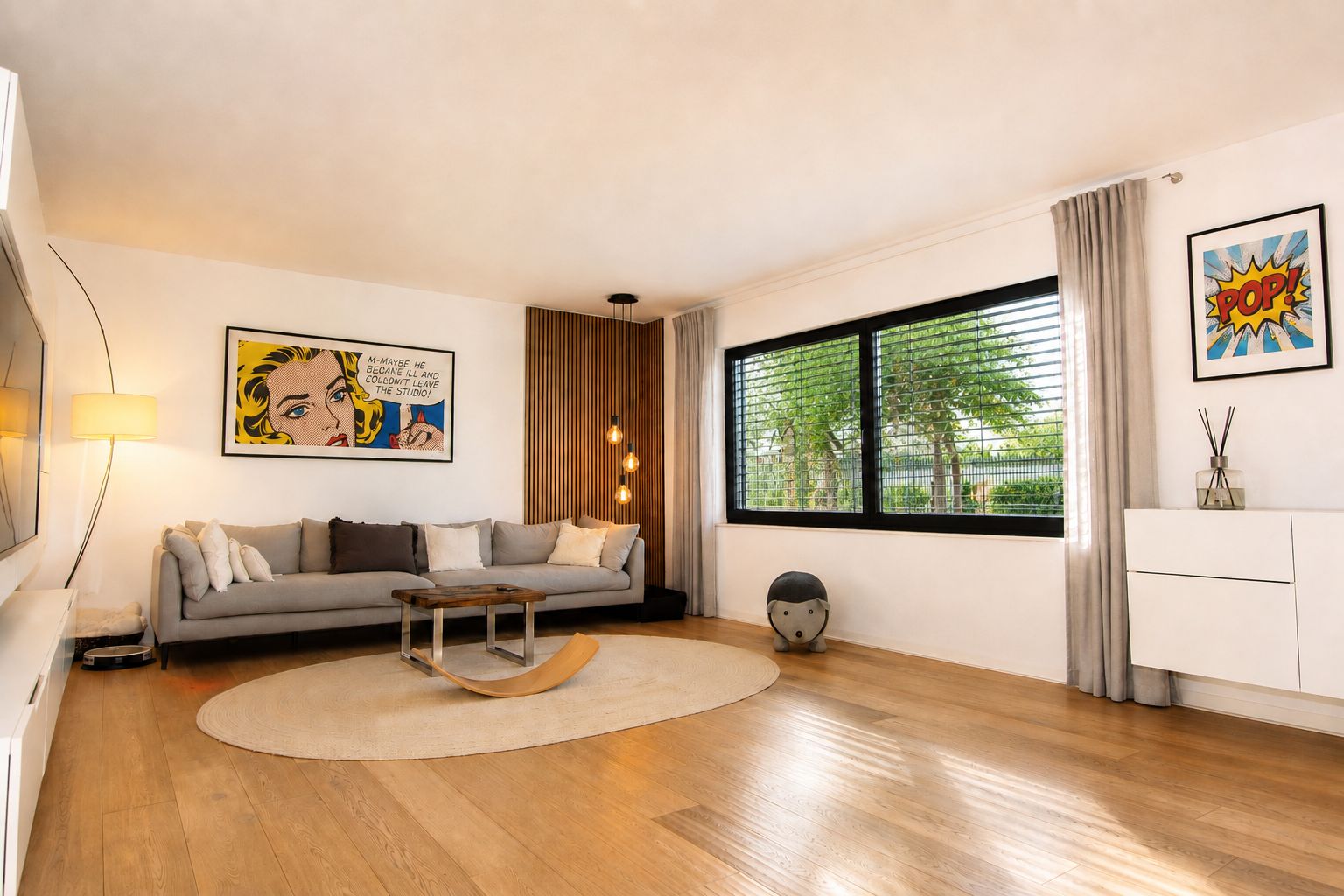 Modernes Wohnzimmer mit grauem Sofa, großem Fenster, rundem Teppich und Pop-Art-Bildern an den Wänden.