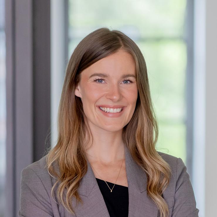 Frau mit langen blonden Haaren im grauen Blazer steht lächelnd mit verschränkten Armen.