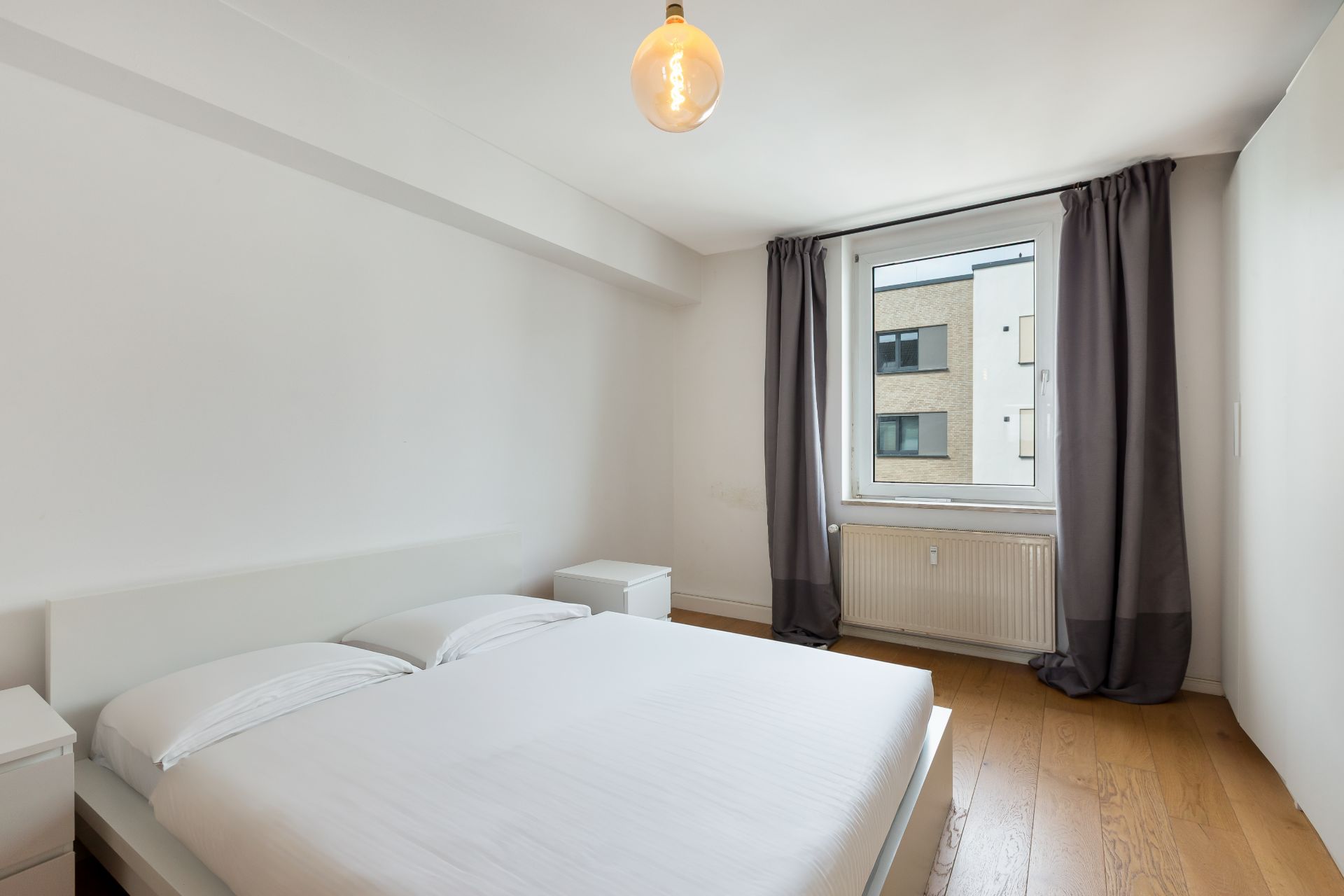 Helles Schlafzimmer mit Doppelbett, Nachttischen, Fenster mit grauen Vorhängen und Holzboden.