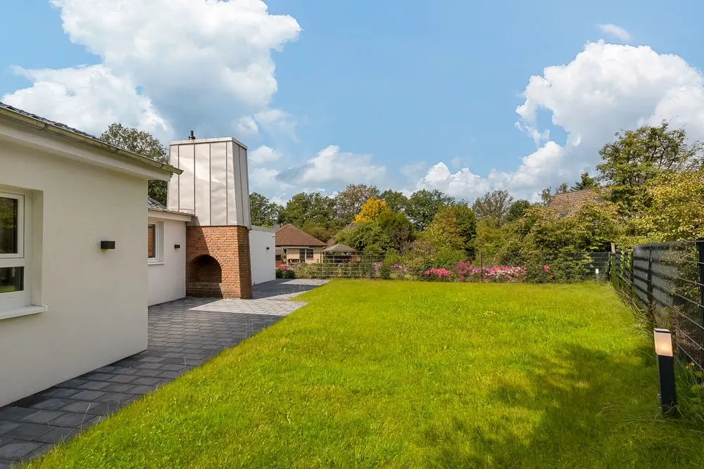 Grüner Garten mit Terrasse und gemauertem Außenkamin neben einem weißen Wohnhaus.