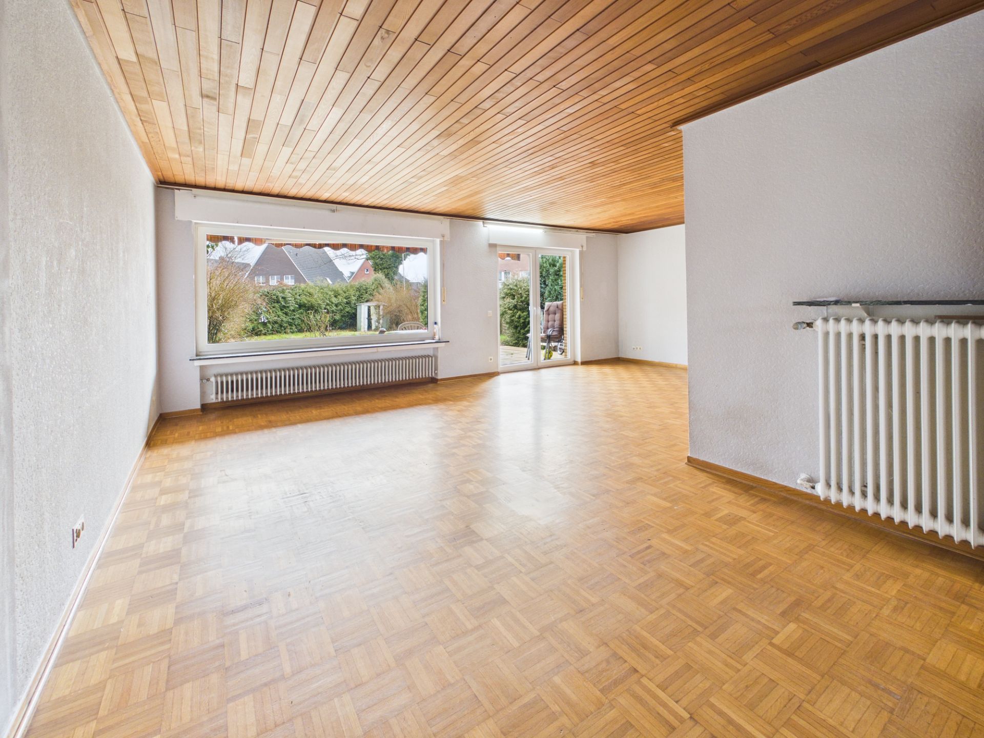 Helles Wohnzimmer mit Parkettboden, Holzdecke, großem Fenster und Heizkörper.