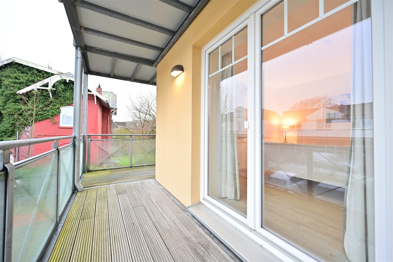 Überdachter Balkon mit Holzfußboden und Glasgeländer, Zugang durch große Glastür.