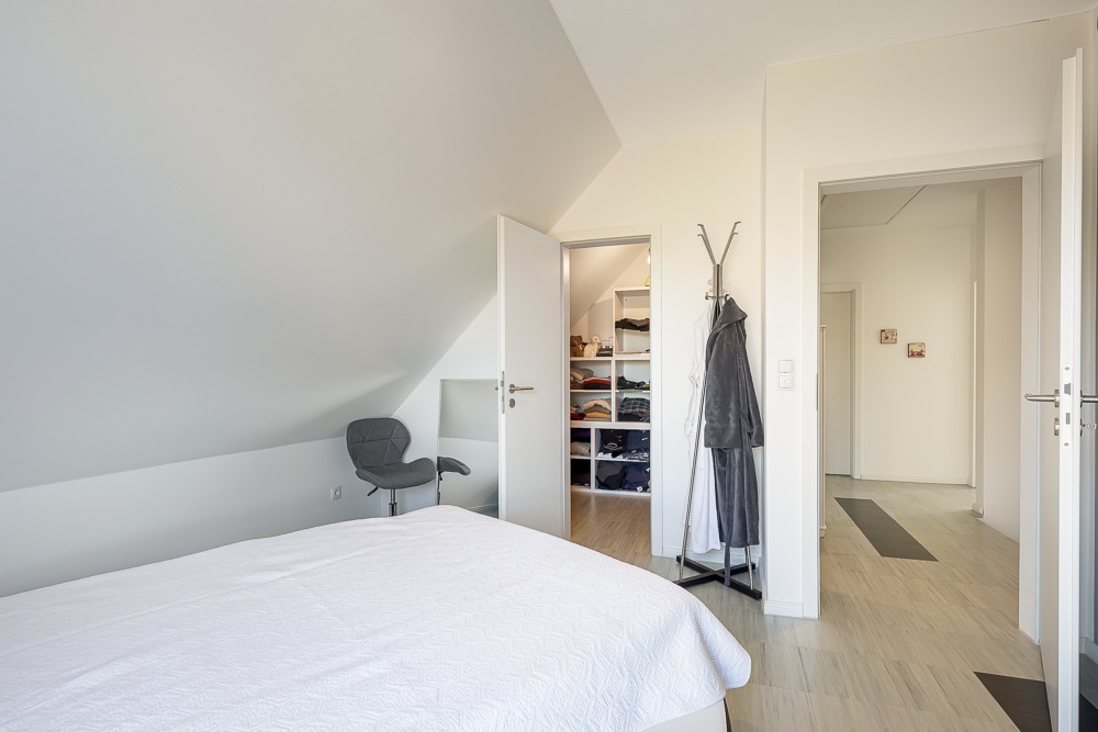 Schlafzimmer mit Bett, Stuhl, offener Tür zum Kleiderschrank und Flur.