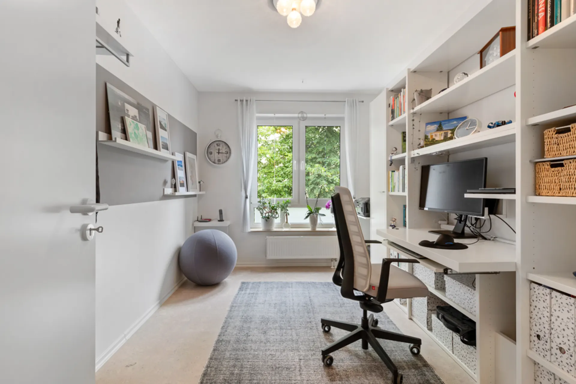 Helles Arbeitszimmer mit Schreibtisch, Regal, Computer, Gymnastikball und großem Fenster mit Blick ins Grüne.