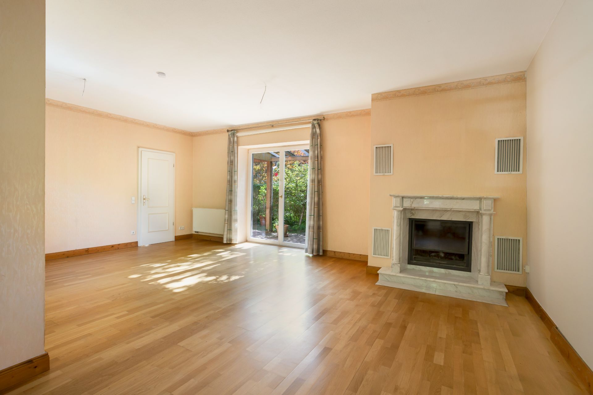 Helles Wohnzimmer mit Kamin, Parkettboden, Glastür und Vorhängen, Blick in den Garten.