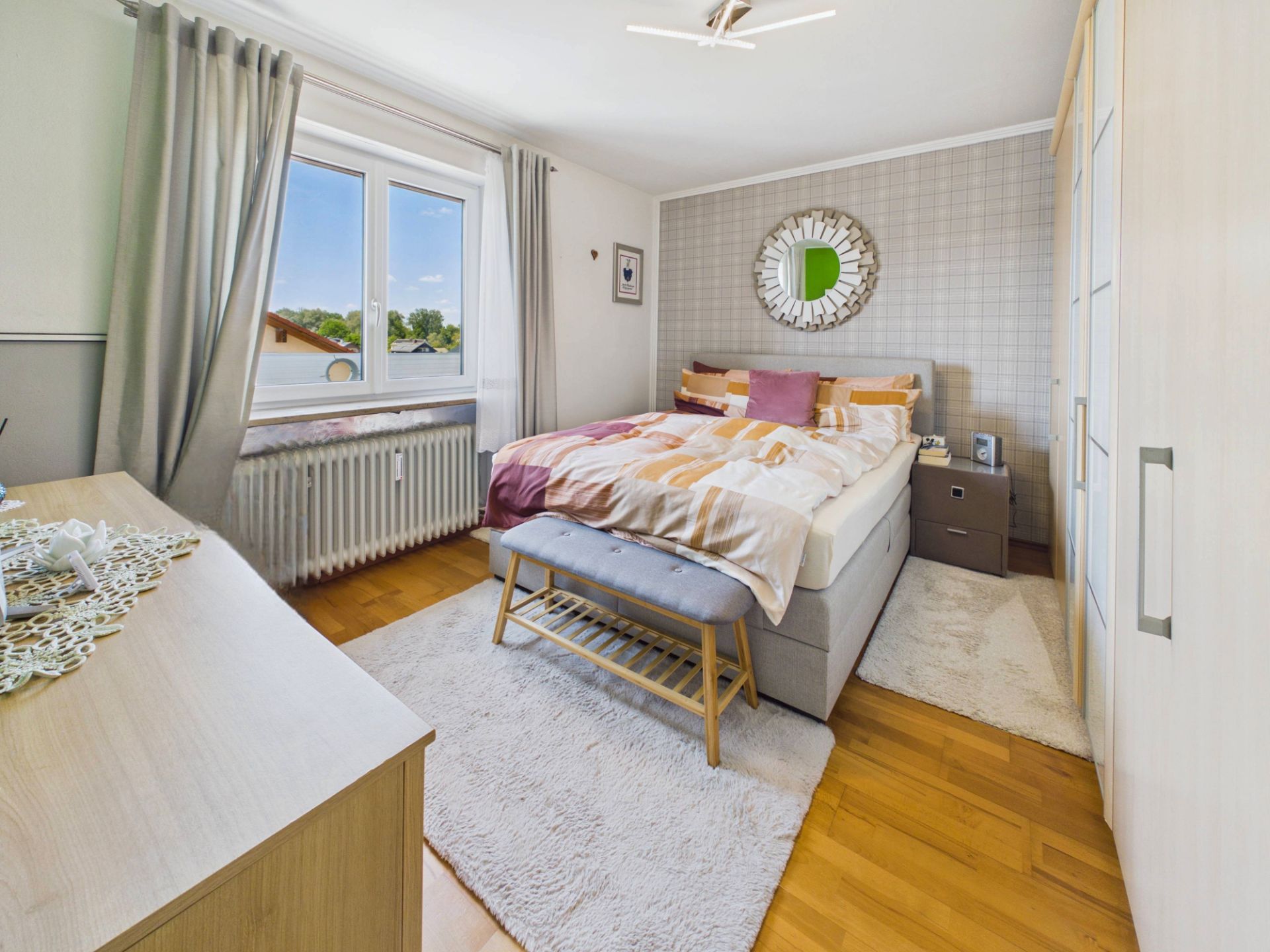 Helles Schlafzimmer mit Doppelbett, großem Fenster, Teppichen und Kleiderschrank auf Holzfußboden.