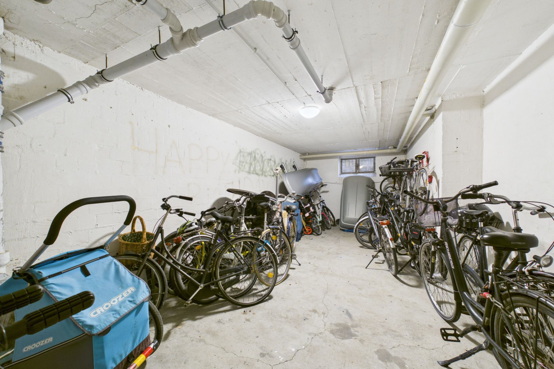 Mehrere Fahrräder stehen in einem Kellerraum mit weißen Wänden und sichtbaren Rohrleitungen.