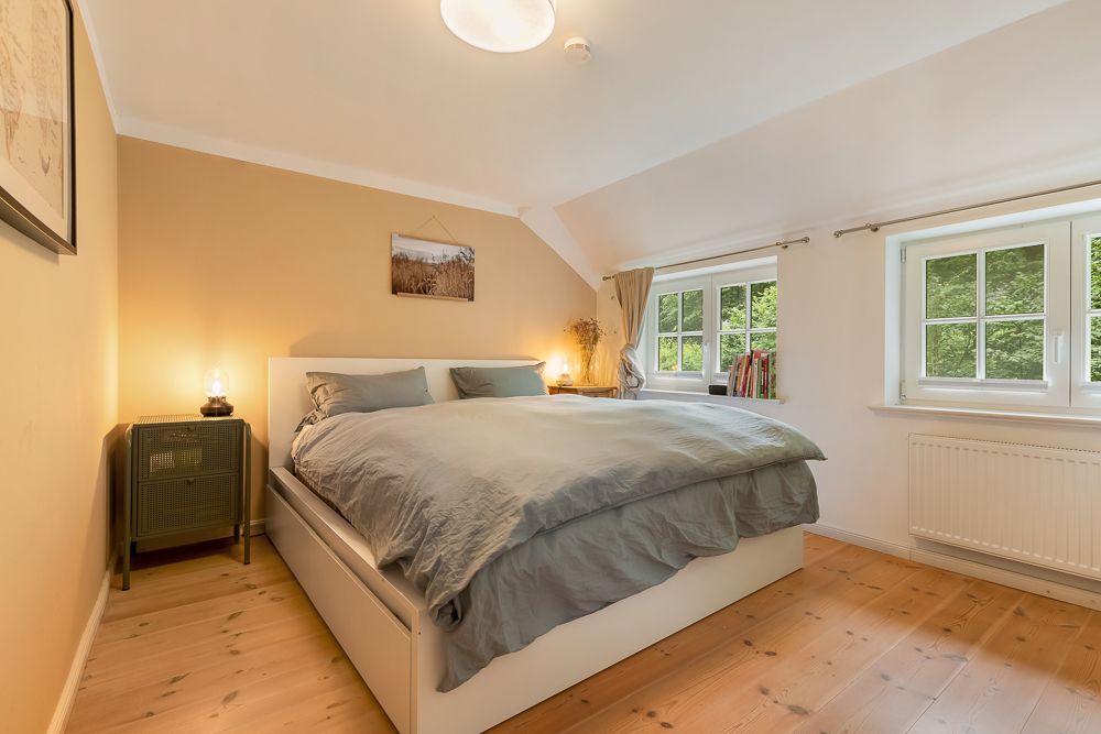 Helles Schlafzimmer mit Doppelbett, Holzboden, Nachttisch und zwei Fenstern.