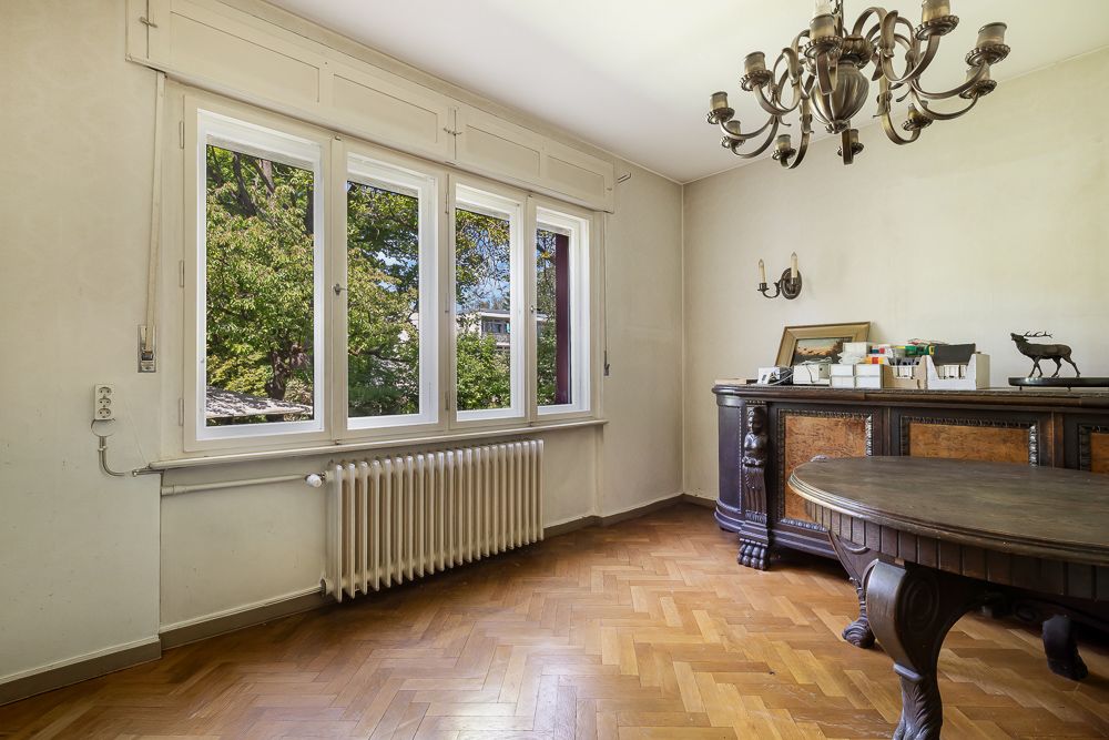 Helles Zimmer mit großem Fenster, Holzfußboden, Heizkörper und dunklem Holztisch mit Kommode.
