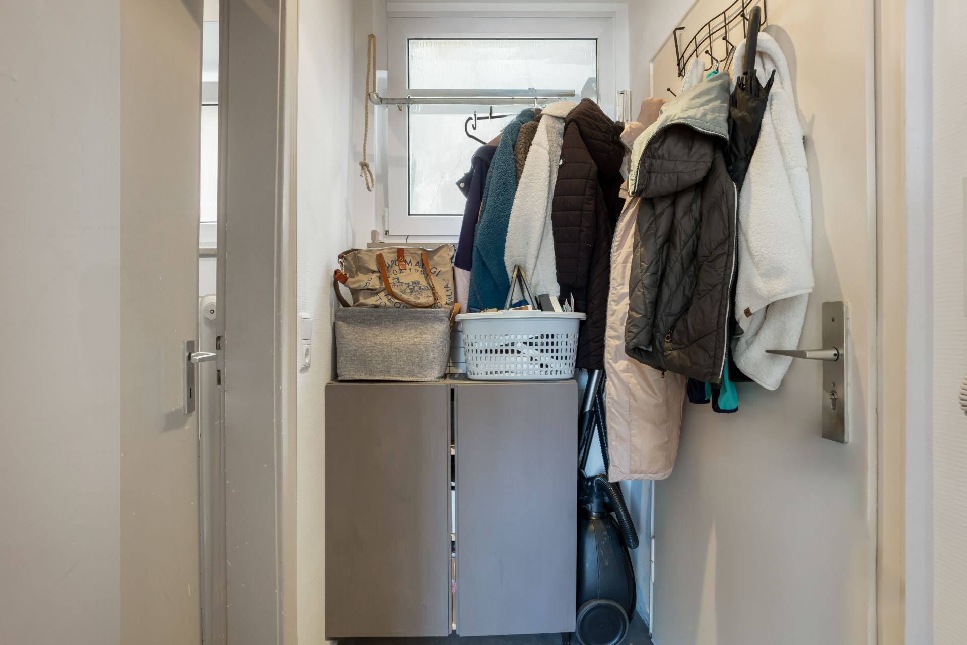 Schmaler Flur mit Schrank, Garderobe, Jacken, Taschen und Haushaltsgegenständen vor einem Fenster.