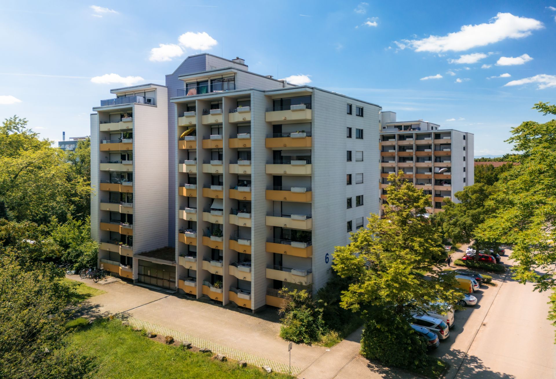 Mehrstöckiges Wohngebäude mit Balkonen und parkenden Autos davor, umgeben von Bäumen.