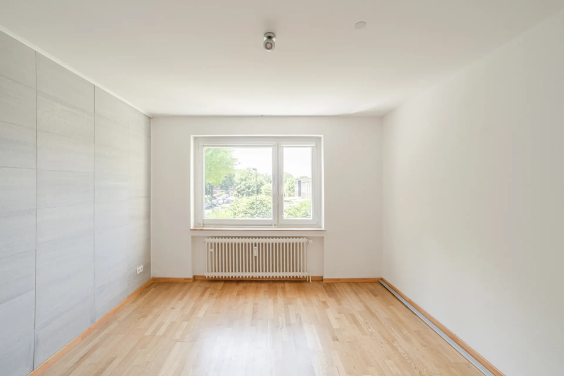 Helles, leeres Zimmer mit Holzboden, großem Fenster und Heizkörper darunter.
