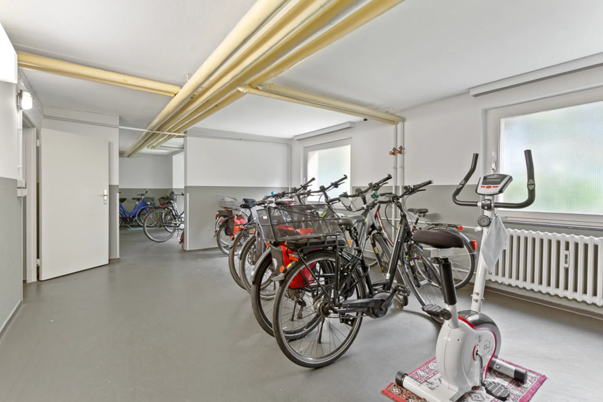 Fahrradraum mit mehreren Fahrrädern und einem Heimtrainer, helle Fenster und offene Tür.