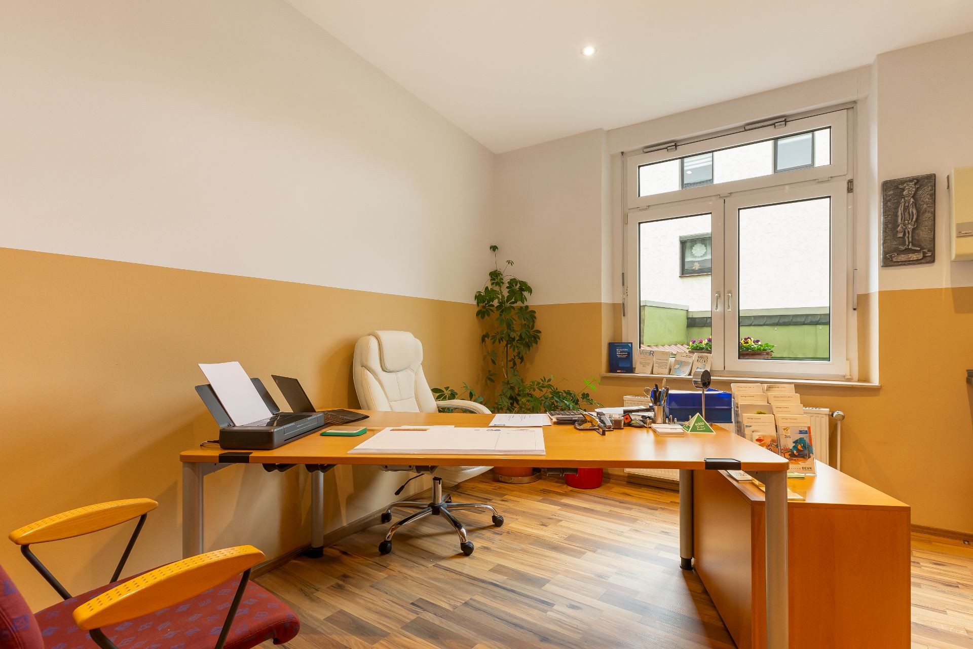Helles Arbeitszimmer mit großem Schreibtisch, Bürostuhl, Pflanzen und Fenster.