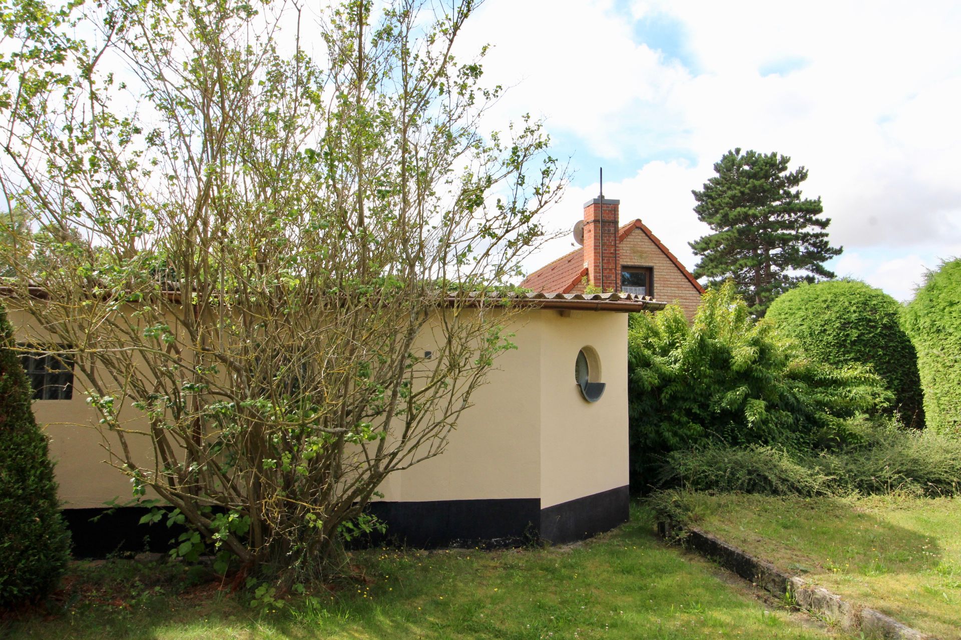 Beigefarbenes Gebäude mit rundem Fenster und Garten mit Sträuchern und Bäumen.