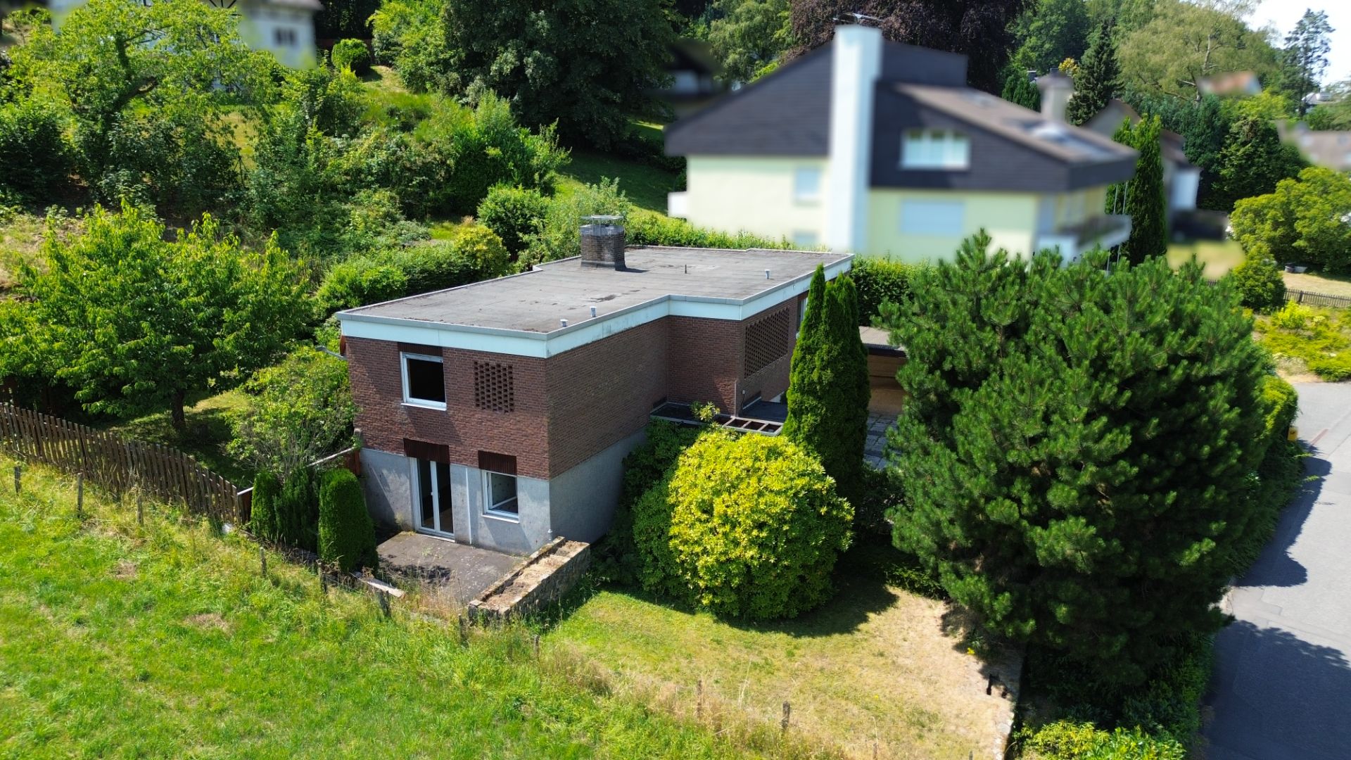 Zweistöckiges Backsteinhaus mit Flachdach, umgeben von Bäumen und Garten.