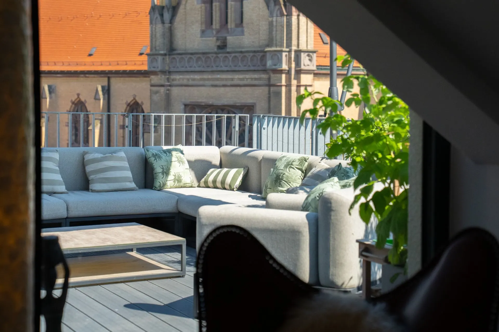 Helle Terrasse mit grauem Ecksofa, Kissen, Couchtisch und Pflanzen, Blick auf ein Gebäude im Hintergrund.