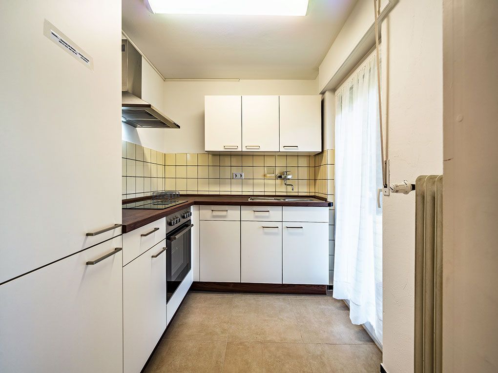 Helle Küche mit weißen Schränken, Herd, Spüle und großem Fenster mit Vorhang.