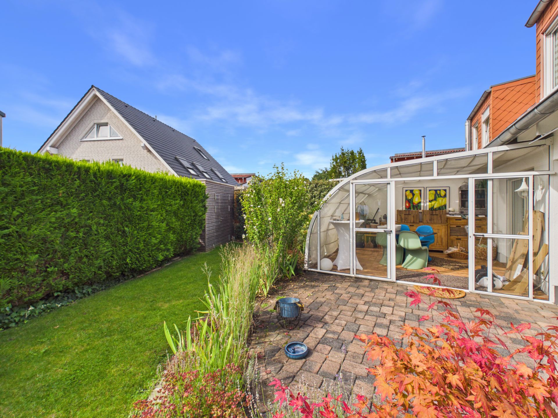 Garten mit Rasen, Terrasse und Wintergarten an einem Wohnhaus bei blauem Himmel.