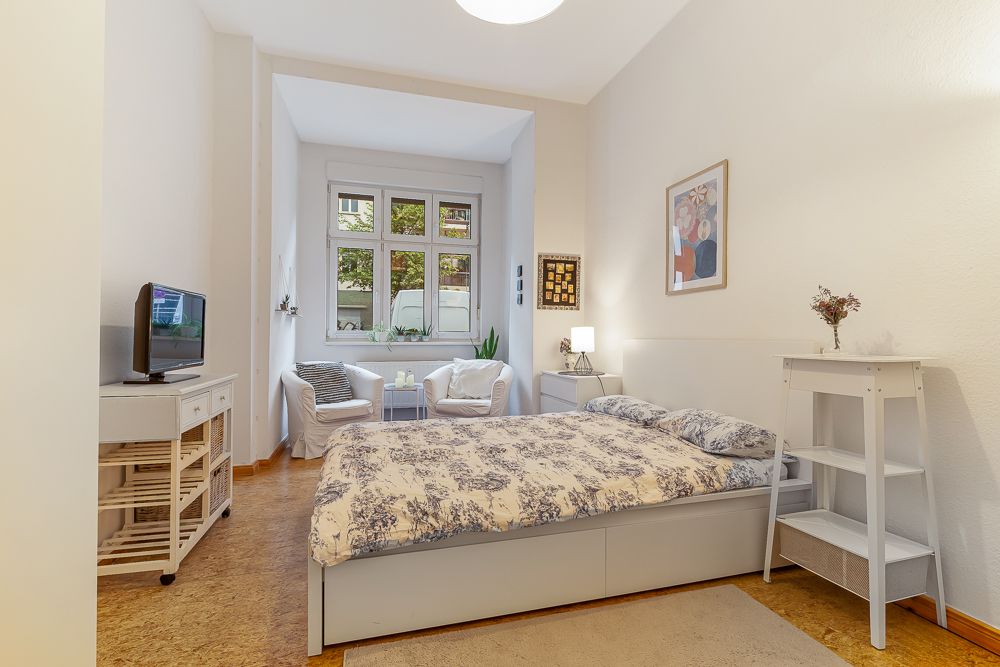 Helles Schlafzimmer mit Doppelbett, Fernseher, zwei Sesseln und großem Fenster.