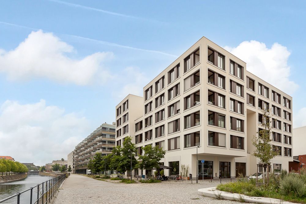 Modernes mehrstöckiges Wohngebäude an einer Uferpromenade mit Bäumen und Gehweg.
