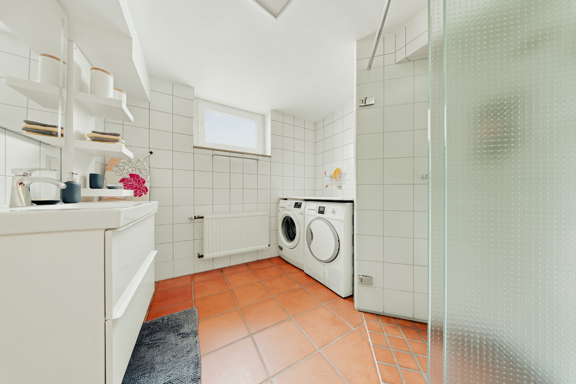 Helles Badezimmer mit Waschbecken, Waschmaschine, Trockner und Dusche mit Glaswand.