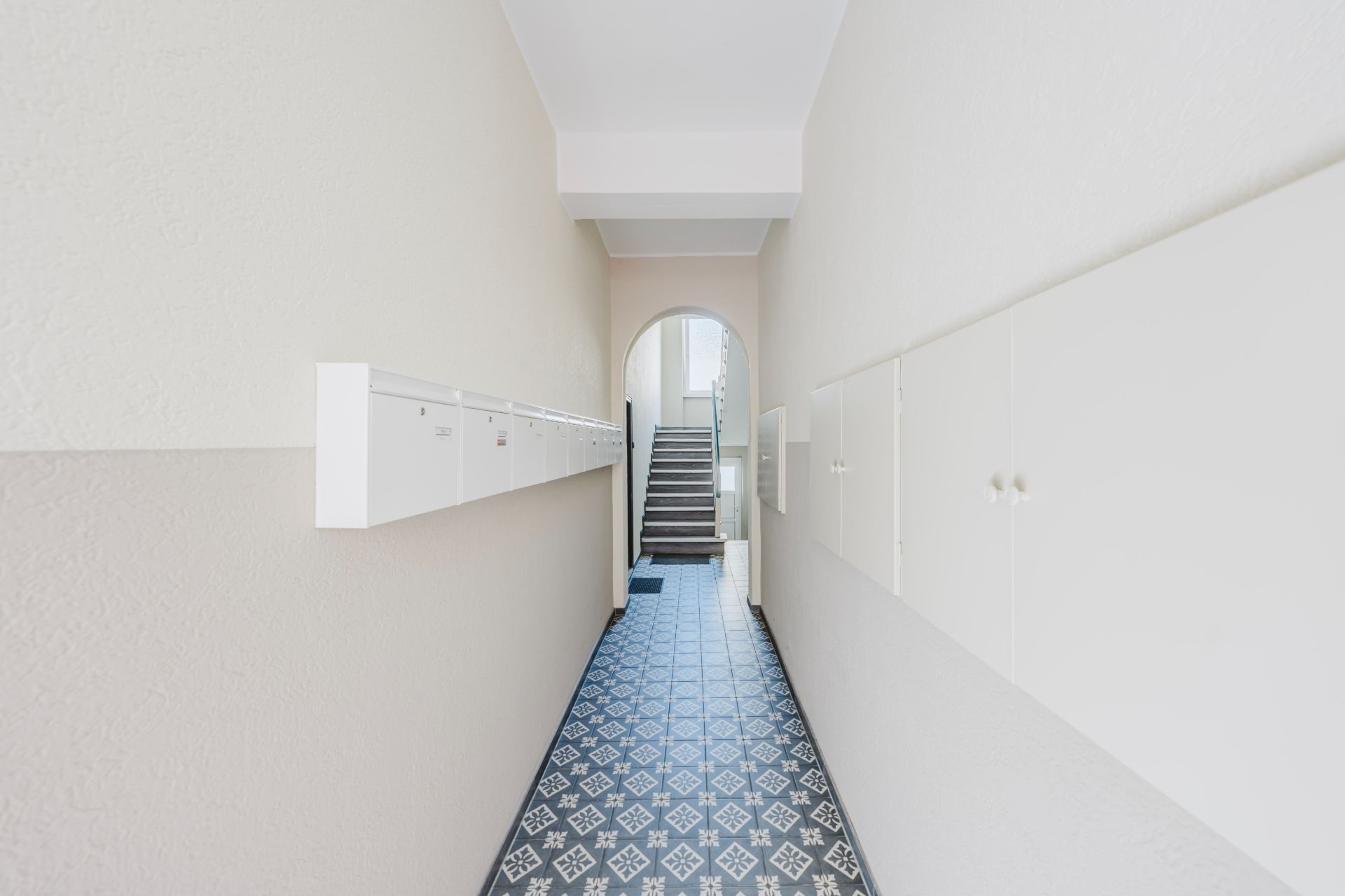 Langer, heller Flur mit Briefkästen an der Wand und gemustertem Boden, am Ende eine Treppe.