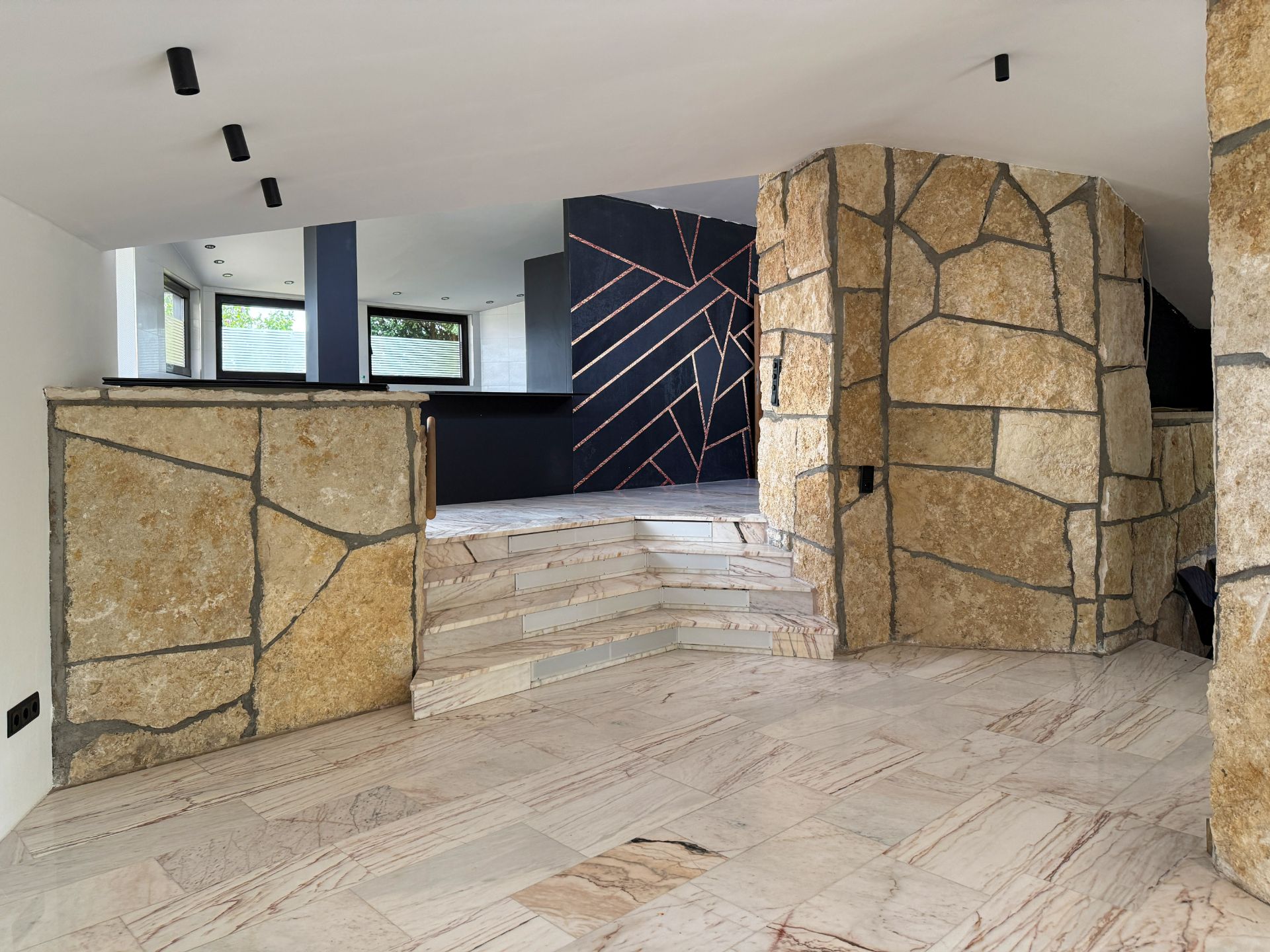 Innenraum mit Natursteinwänden, Marmorboden und Treppe zu einem modernen Bereich mit großen Fenstern.