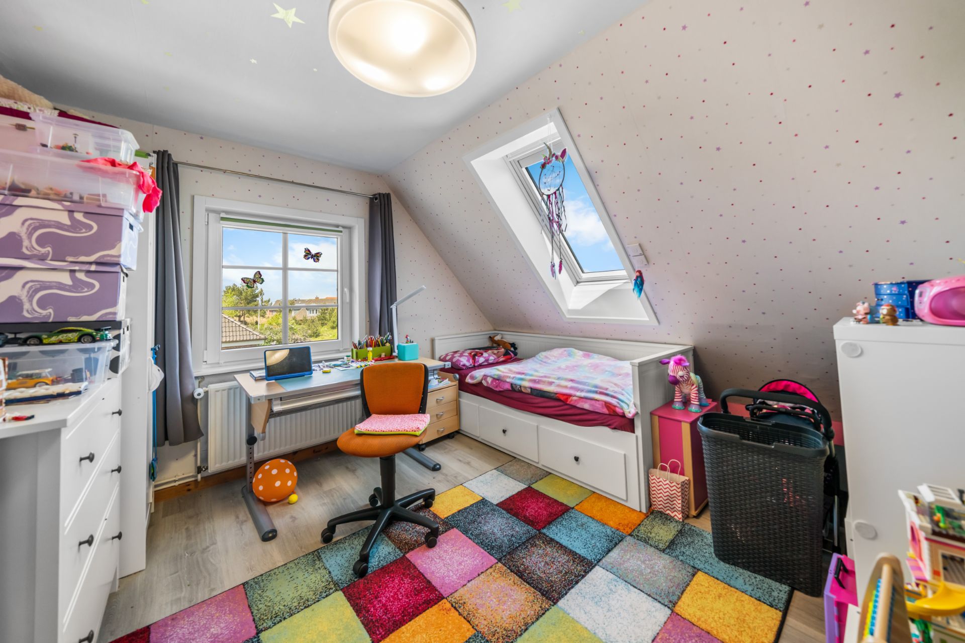 Kinderzimmer mit buntem Teppich, Bett, Schreibtisch und Stuhl unter Dachschräge, Fenster mit Aussicht.