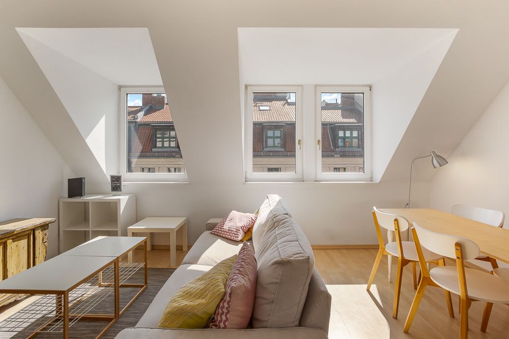 Helles Wohnzimmer mit Sofa, Esstisch, Stühlen und großen Fenstern in einer Dachgeschosswohnung.