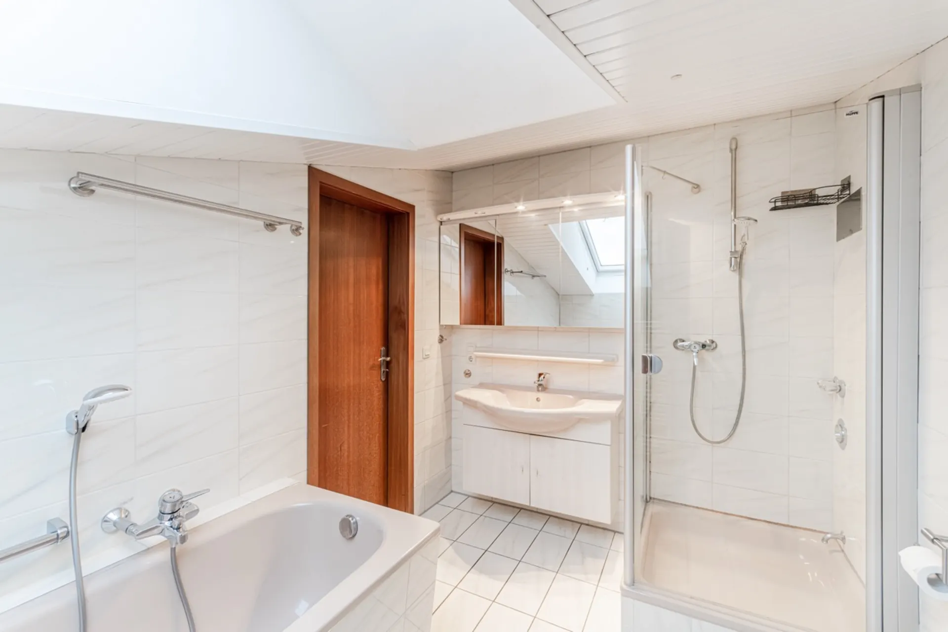 Helles Badezimmer mit Badewanne, Dusche, Waschbecken, Spiegel und Holzinnentür.