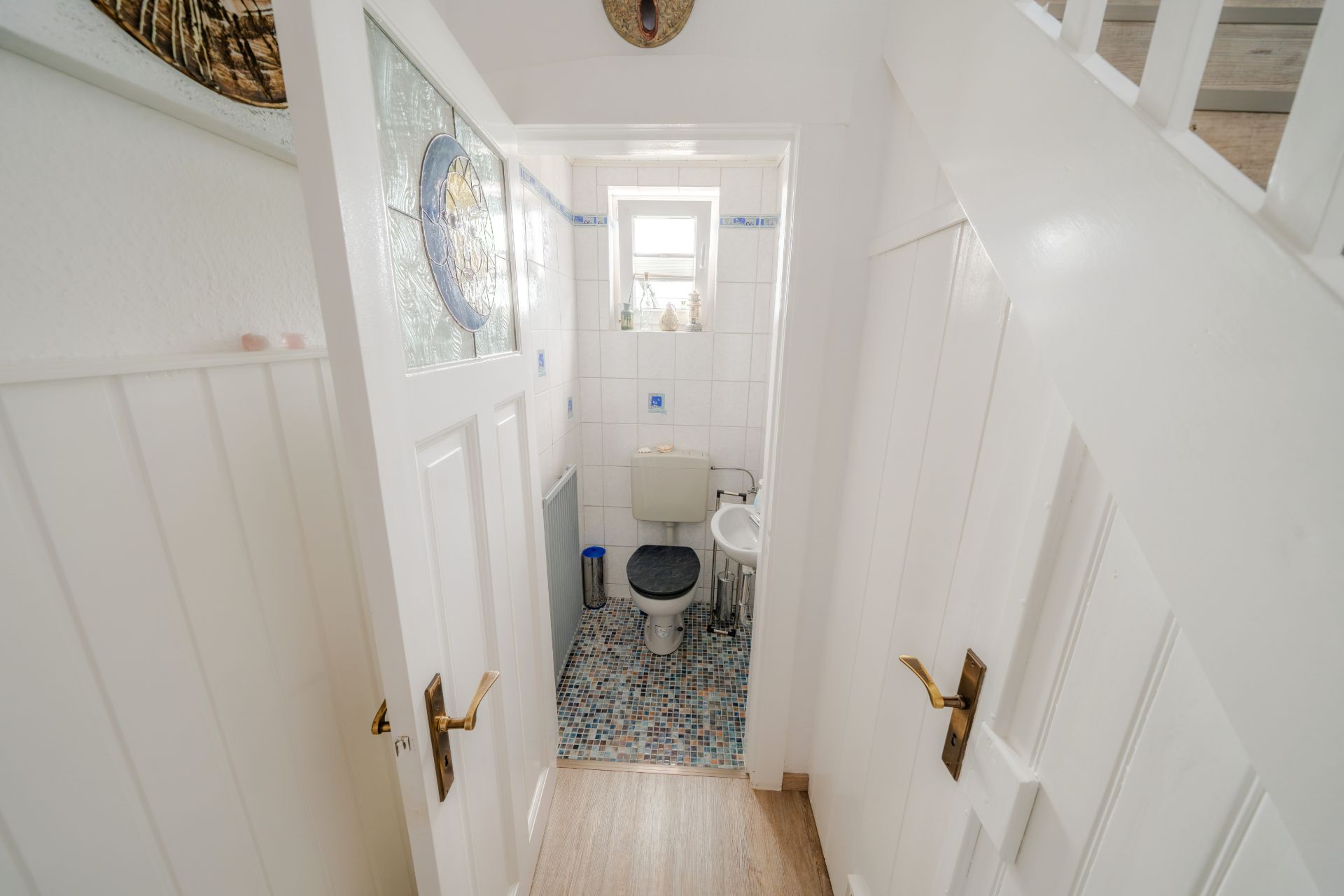 Kleines Bad mit Toilette, Waschbecken und Fenster, weiße Fliesen und Mosaikfliesenboden.