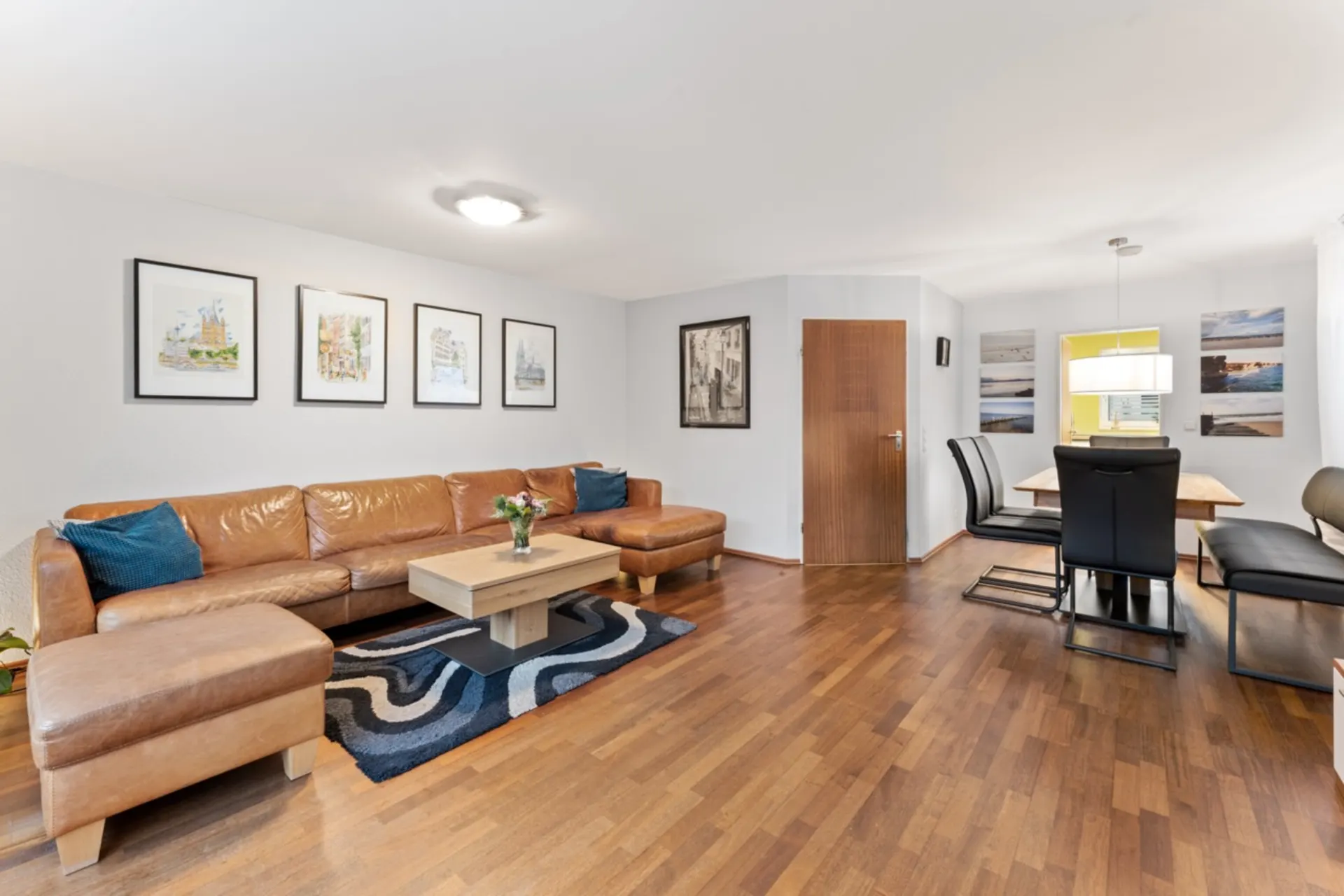 Wohnzimmer mit braunem Ledersofa, Holztisch und Essbereich mit schwarzen Stühlen auf Parkettboden.