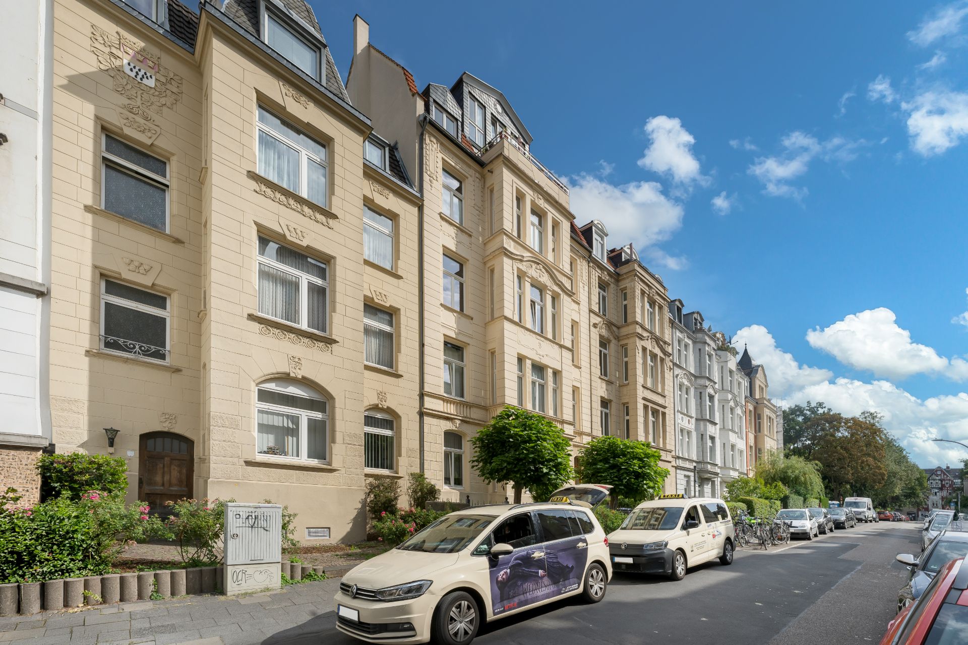 Mehrstöckige Wohngebäude an einer ruhigen Straße mit geparkten Autos und Bäumen vor den Häusern.
