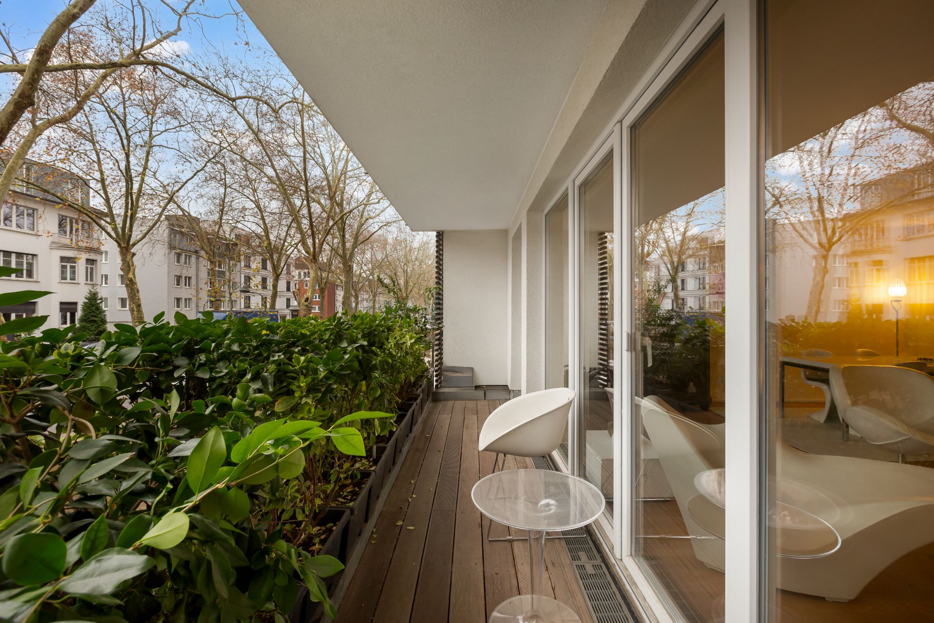 Balkon mit Holzfußboden, Glasgeländer, weißen Stühlen und Blick auf grüne Pflanzen sowie Nachbarhäuser.